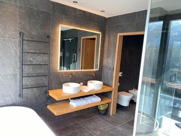 A modern bathroom with dark tiled walls, a large mirror, two sinks, a shower, and a glass-enclosed shower area.