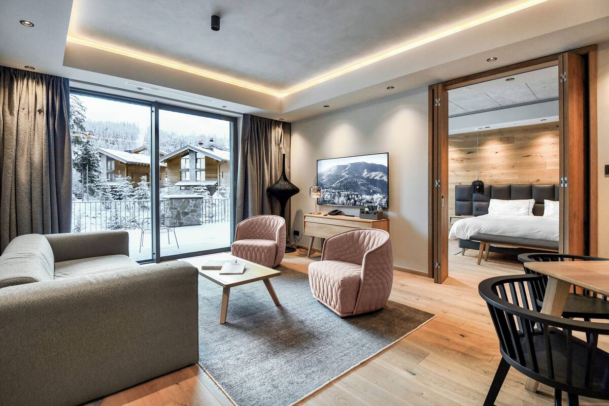 A modern hotel room with a large sliding glass door leading to a balcony, a cozy seating area with a sofa and chairs, a wooden desk with a TV, and a separate bedroom with a bed and a nightstand.