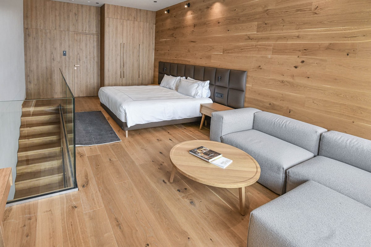 A modern hotel room with a wooden floor, a bed with white linens, a grey sofa, a round wooden coffee table, and a glass railing beside a staircase.