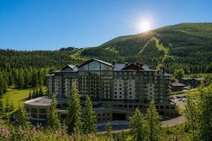A large hotel with multiple floors, surrounded by a forested area and a mountain in the background.