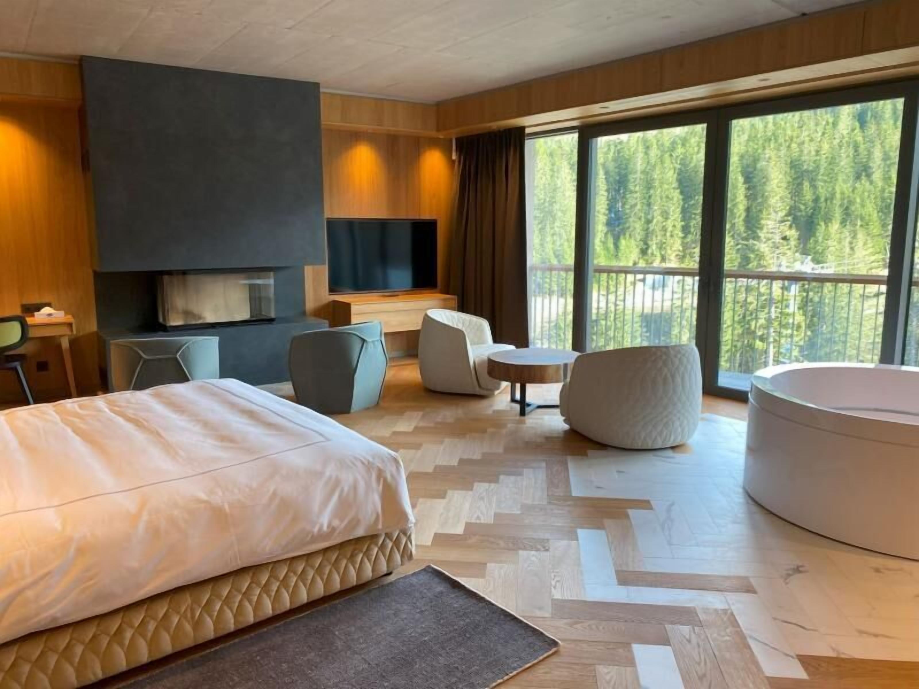 Et moderne hotelværelse med en stor seng, et opholdsområde med stole og et bord, et fjernsyn og et stort vindue med udsigt til et skovområde.