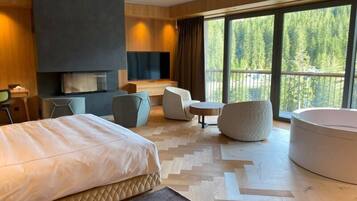 Et moderne hotelværelse med en stor seng, et opholdsområde med stole og et bord, et fjernsyn og et stort vindue med udsigt til et skovområde.