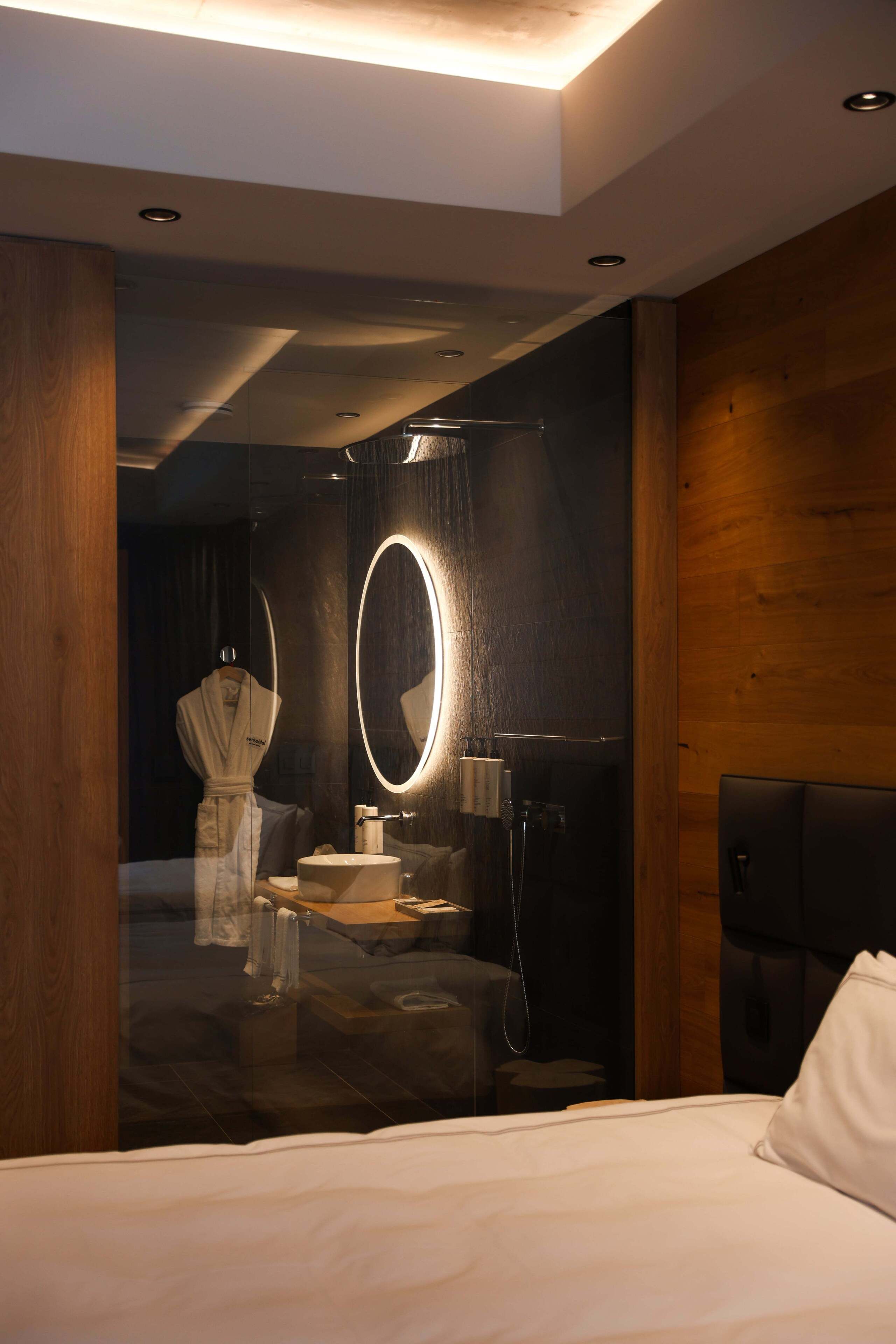 Una habitación de hotel moderna con una cama grande, un espejo redondo, un panel de pared de madera y una pequeña zona de estar con una mesa y una silla.