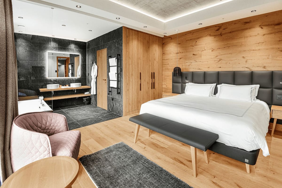 A modern bedroom with a large bed, a wooden headboard, a bedside table, a bench, a chair, a mirror, a vanity with a sink, and a wardrobe.