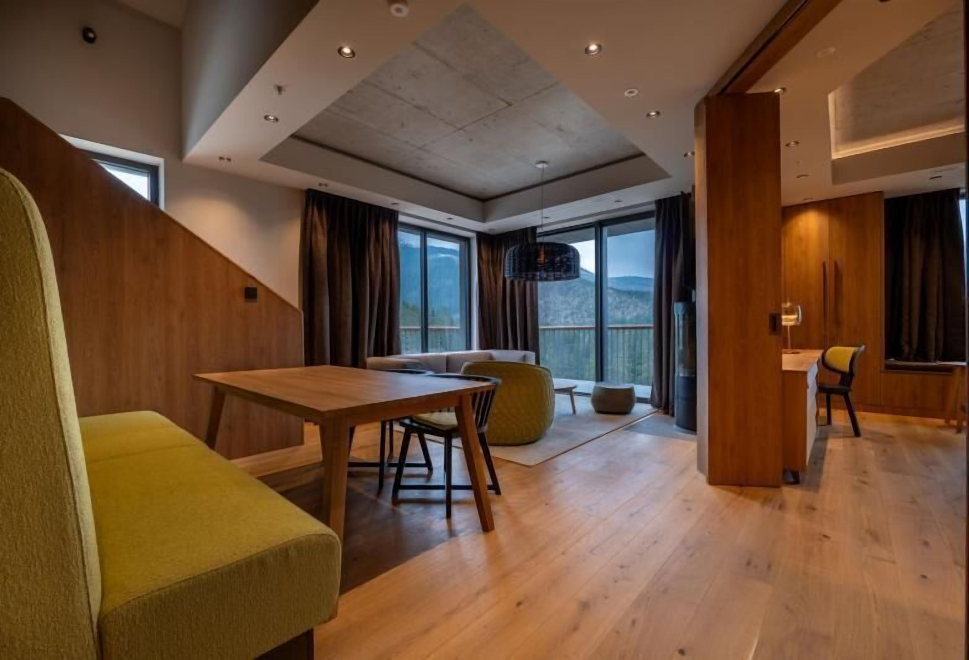 Un intérieur moderne avec une table à manger en bois, un canapé et une chaise. De grandes fenêtres sont agrémentées de rideaux, et on y trouve un luminaire suspendu ainsi qu’un mur d’accent en bois.