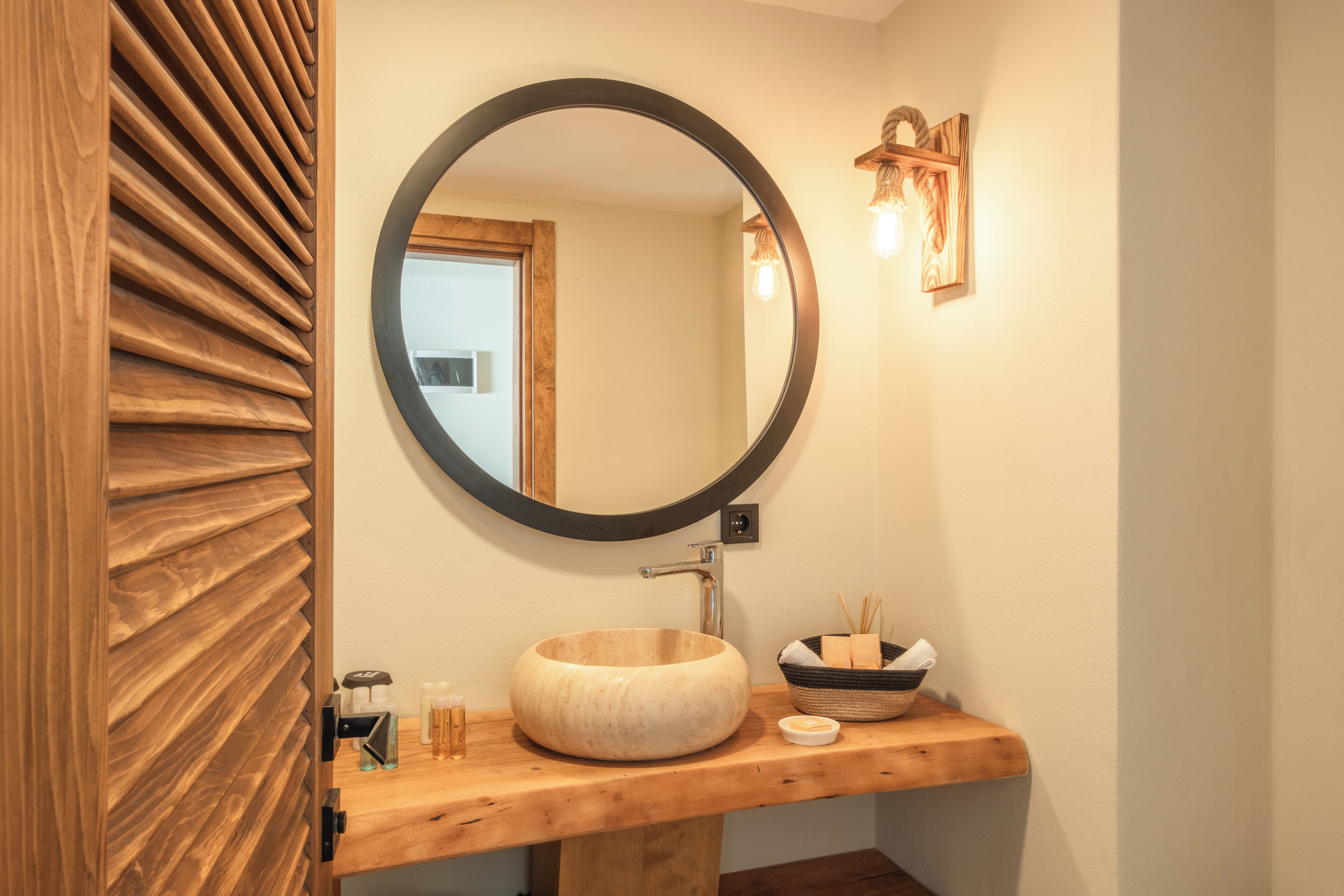 A wooden vanity with a round mirror, a wooden bowl sink, and a small shelf with toiletries.