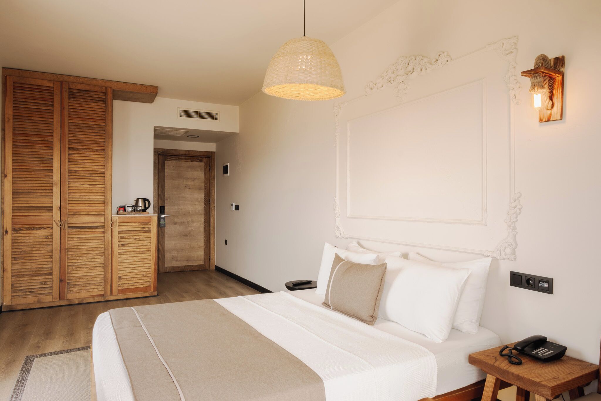 A hotel room with a large bed, wooden wardrobe, a hanging light fixture, and a wall-mounted phone.