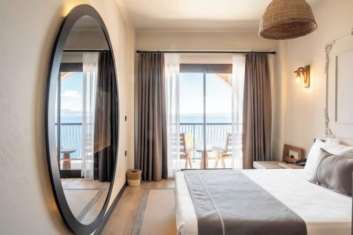 A hotel room with a large bed, a round mirror, a balcony with a view, and a woven light fixture.