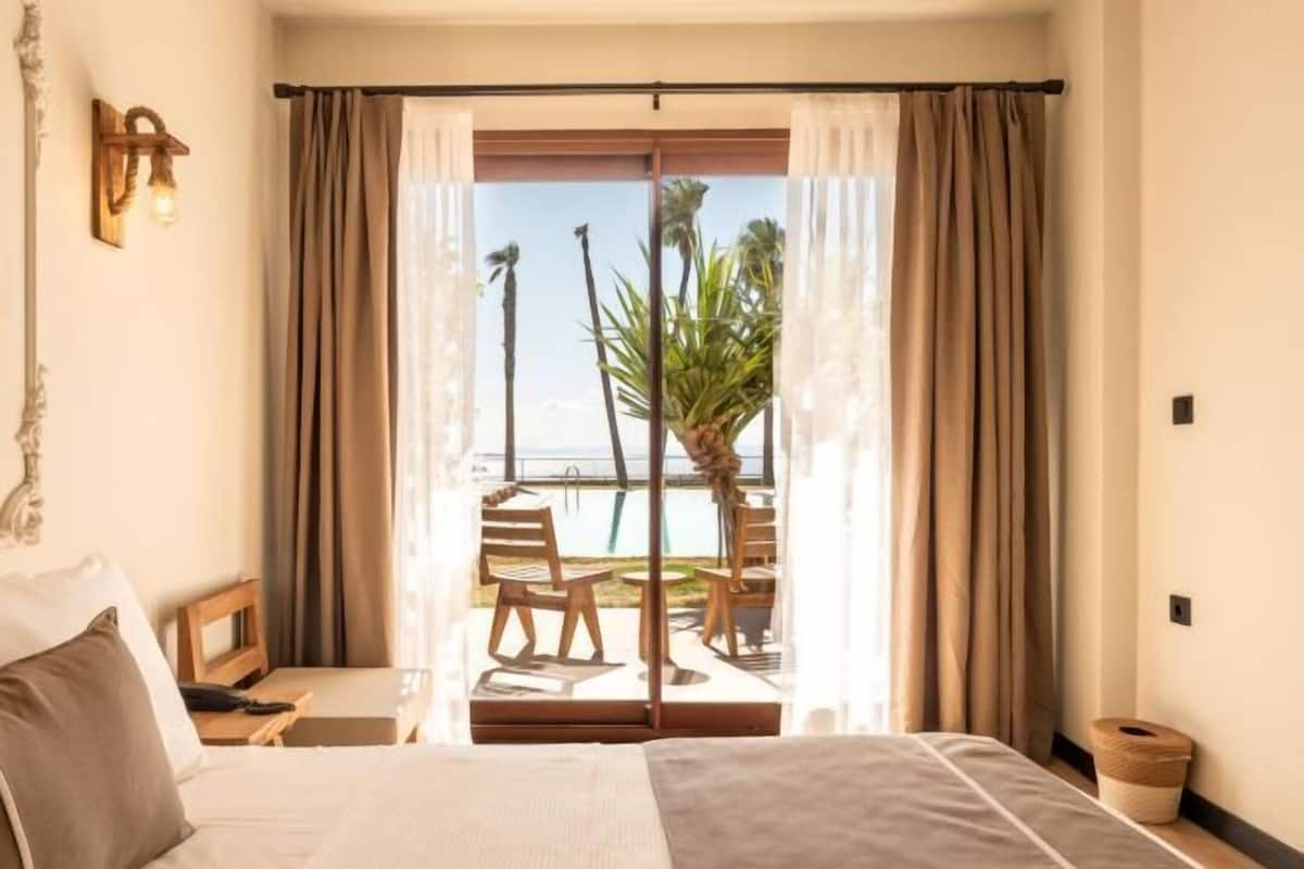 A hotel room with a bed, a desk, a chair, and a view of a pool and palm trees.