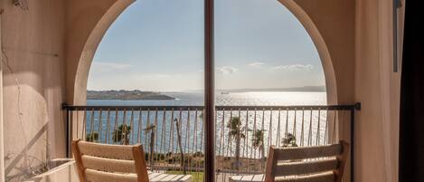 Un balcón con dos sillas de madera y una mesa, con vista a una playa con palmeras y cielo despejado.
