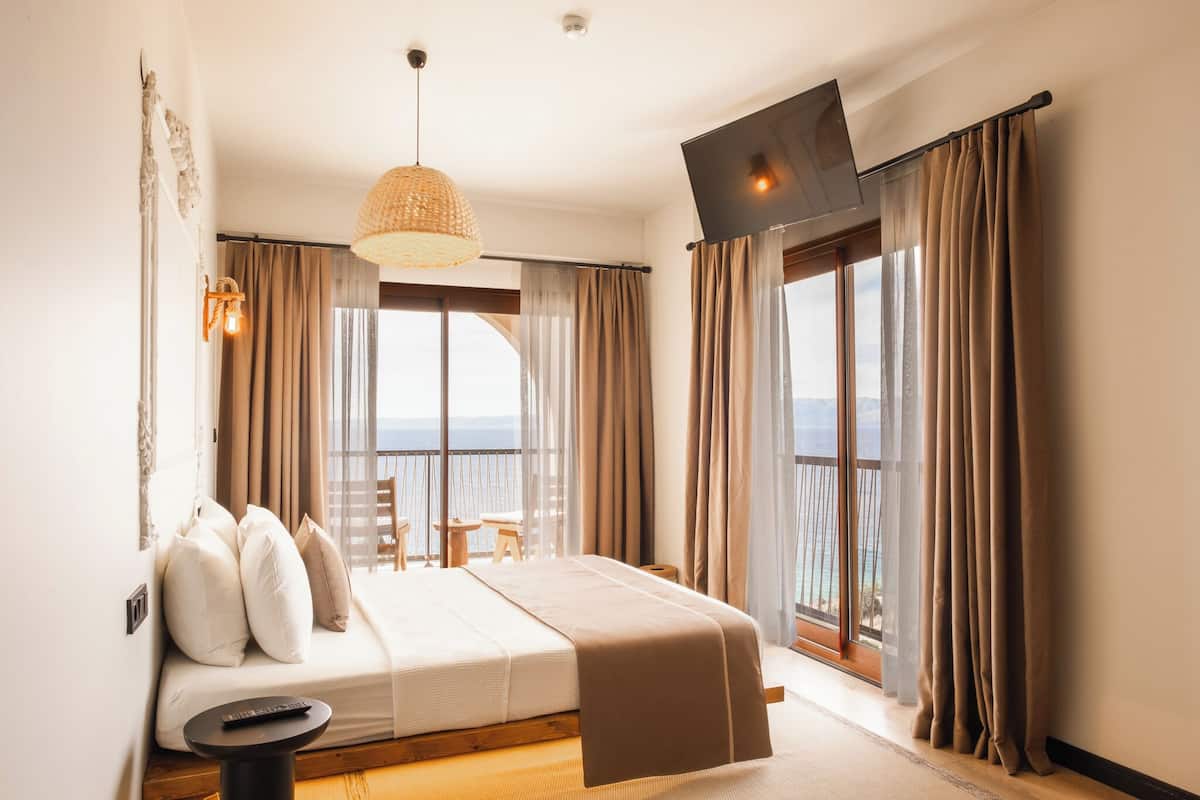 A bedroom with a bed, a TV, a balcony with curtains, and a view of the sea.