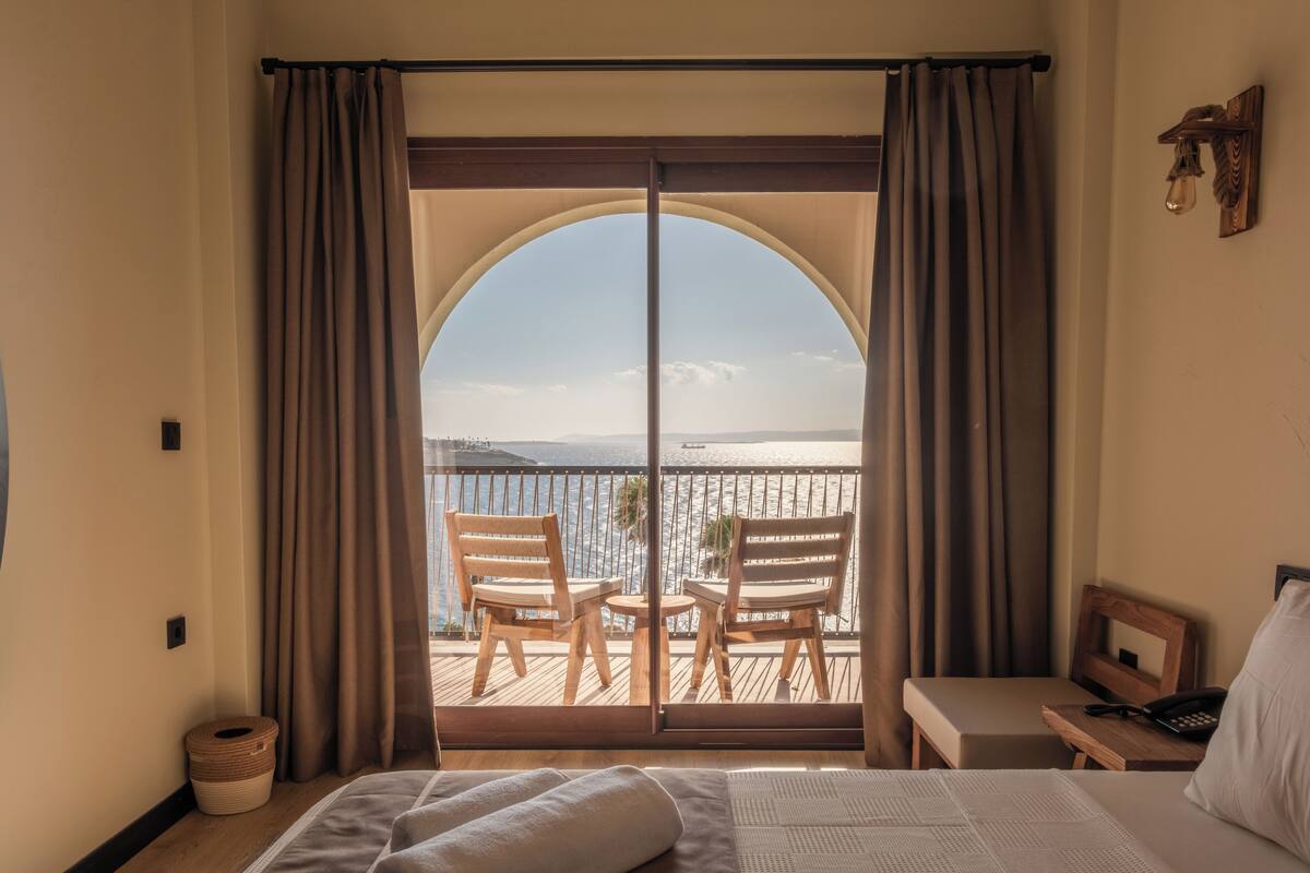 A hotel room with a bed, a desk, a chair, and a view of the sea through a large arched window.