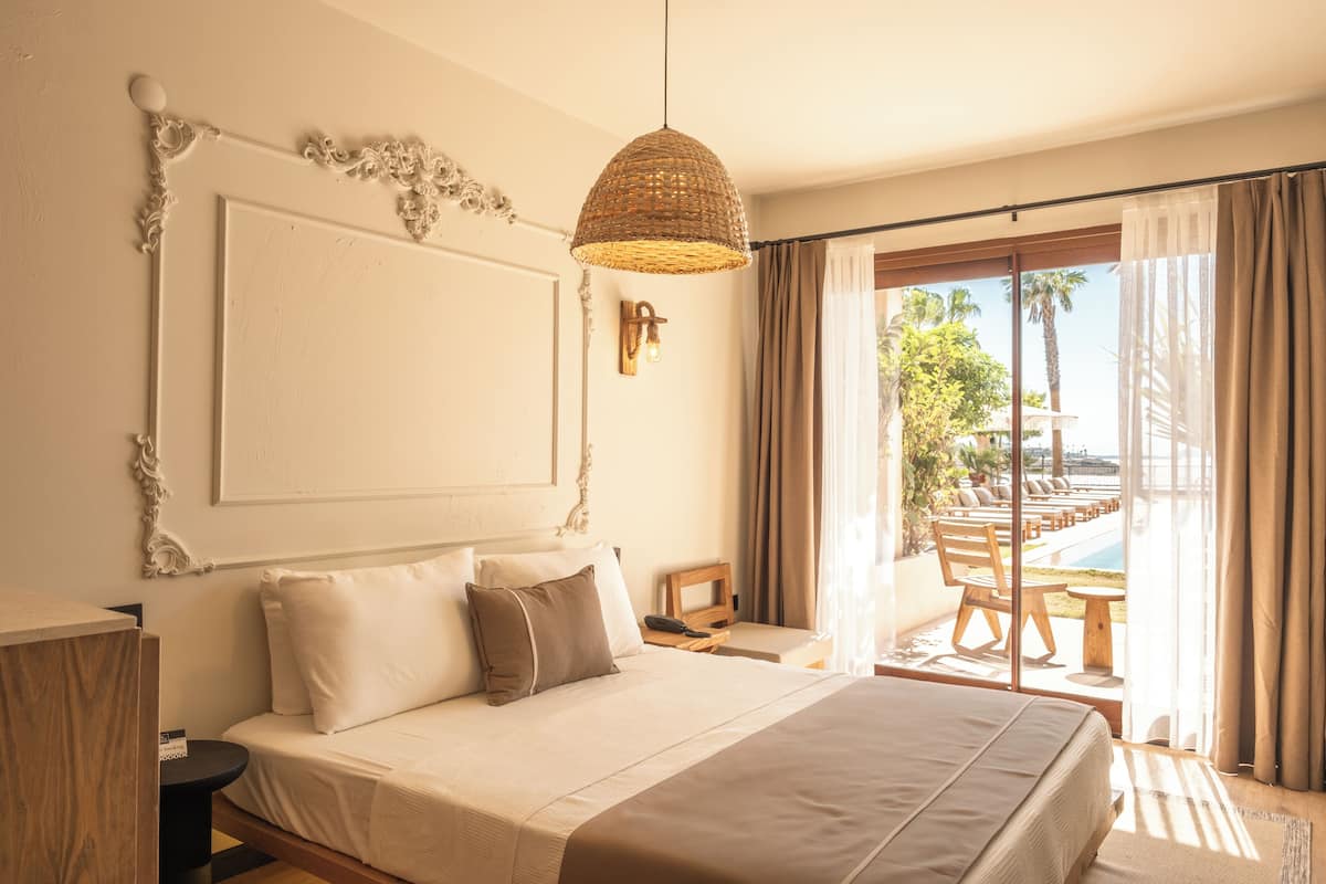 A bedroom with a large bed, a hanging light fixture, a view of a pool and palm trees through the window.