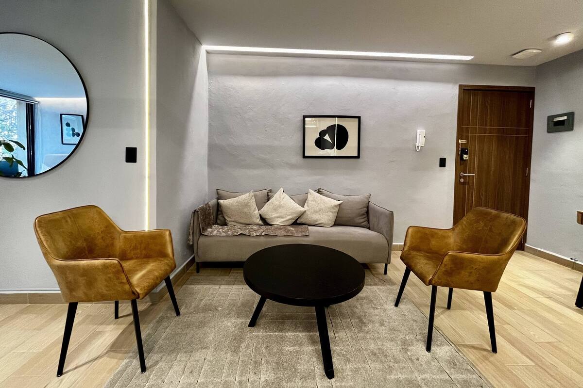 Ein modernes Wohnzimmer mit einer Couch, zwei Sesseln, einem Couchtisch, einem runden Spiegel und einem gerahmten abstrakten Kunstwerk an der Wand.