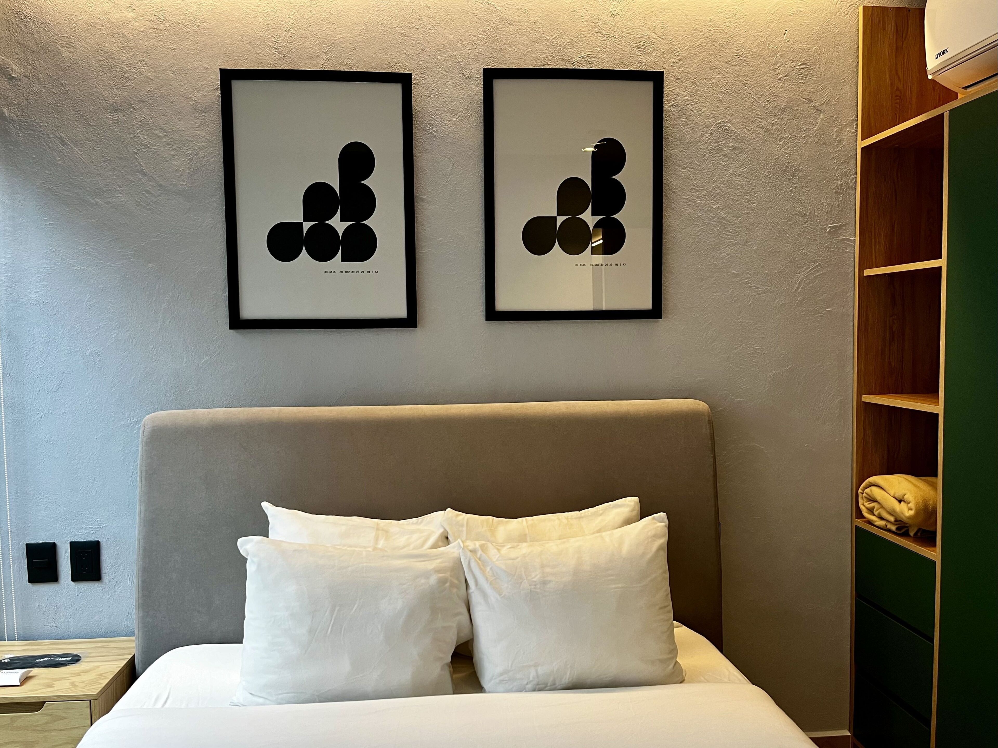A hotel room with a bed, two framed abstract artworks, a bedside table, and a green storage unit.