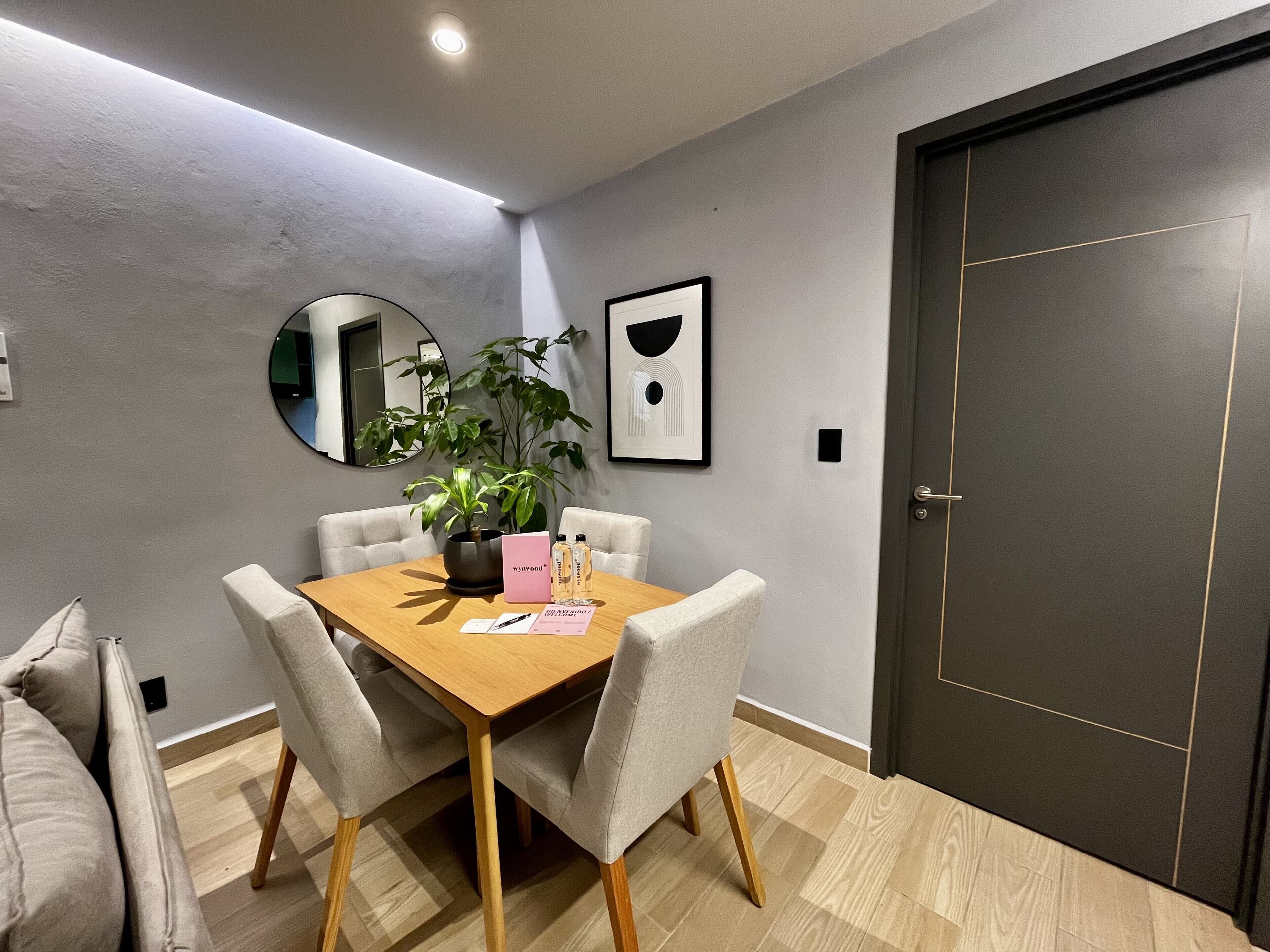 A modern dining area with a wooden table, four chairs, a potted plant, and a framed artwork on the wall.