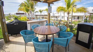 Una terrazza esterna con un tavolo rotondo in legno e sedie in vimini blu, circondata da palme e vista su una zona residenziale.