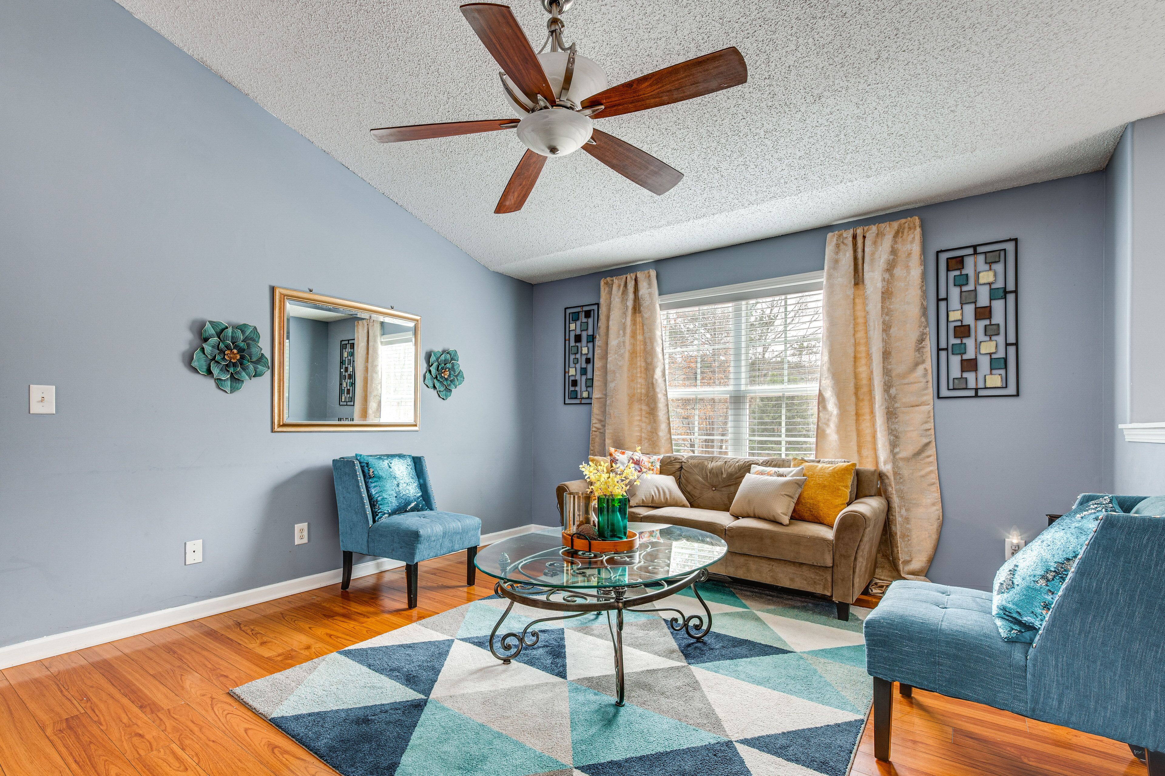 A living room with a ceiling fan, a sofa, two armchairs, a glass coffee table, and a patterned rug.