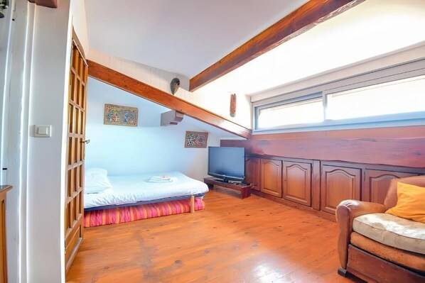 A bedroom with a bed, a television, wooden flooring, a window, and a wooden ceiling beam.