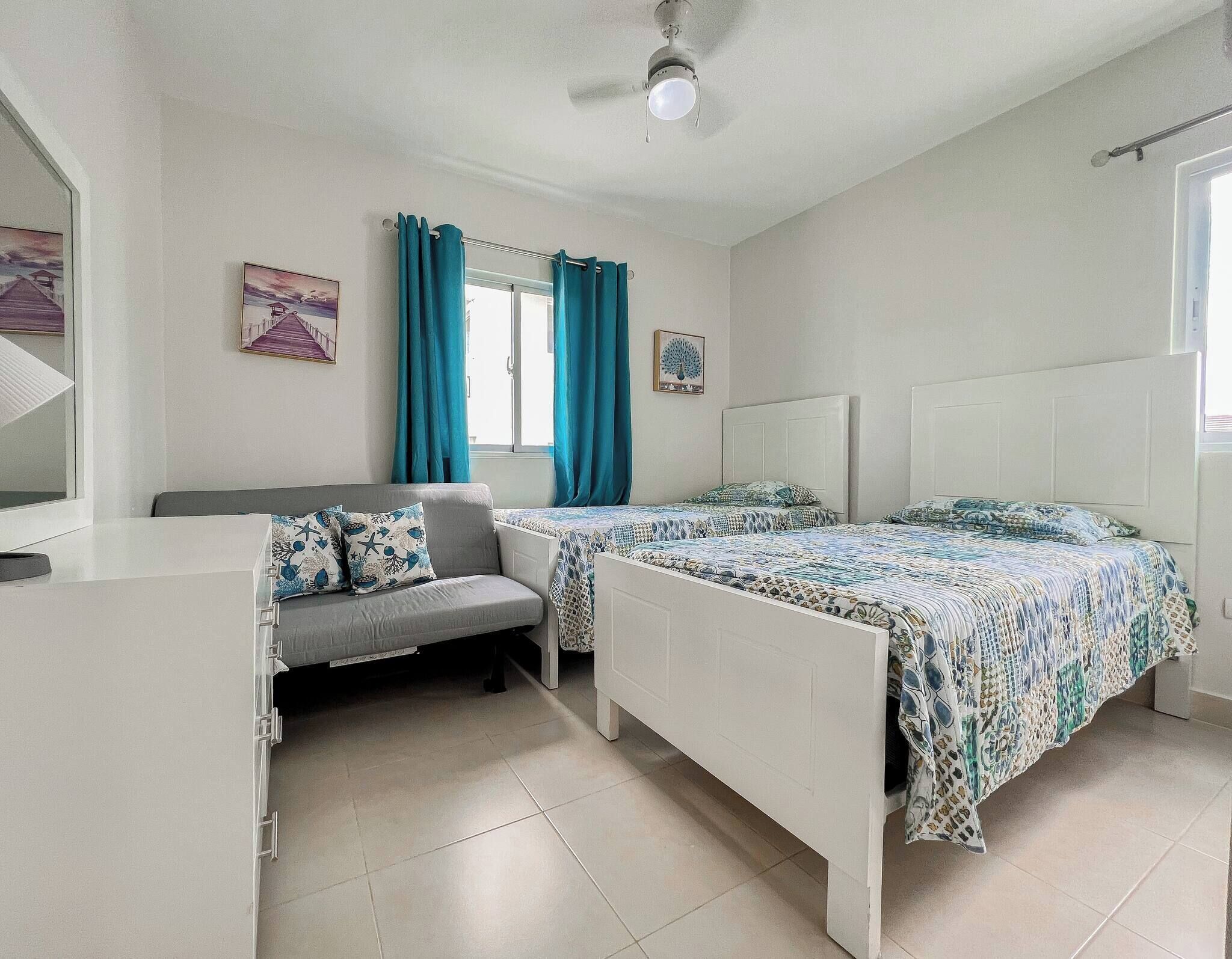 A bedroom with two beds, a sofa, a window with curtains, and a ceiling fan.