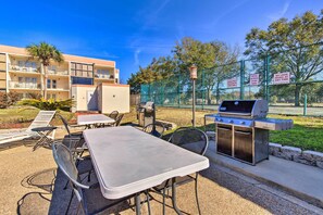 An outdoor patio with a grill, tables, and chairs, surrounded by a fence and a building in the background. - Biloxi Condo w/ Pool Access - Steps to Beach! (Biloxi)