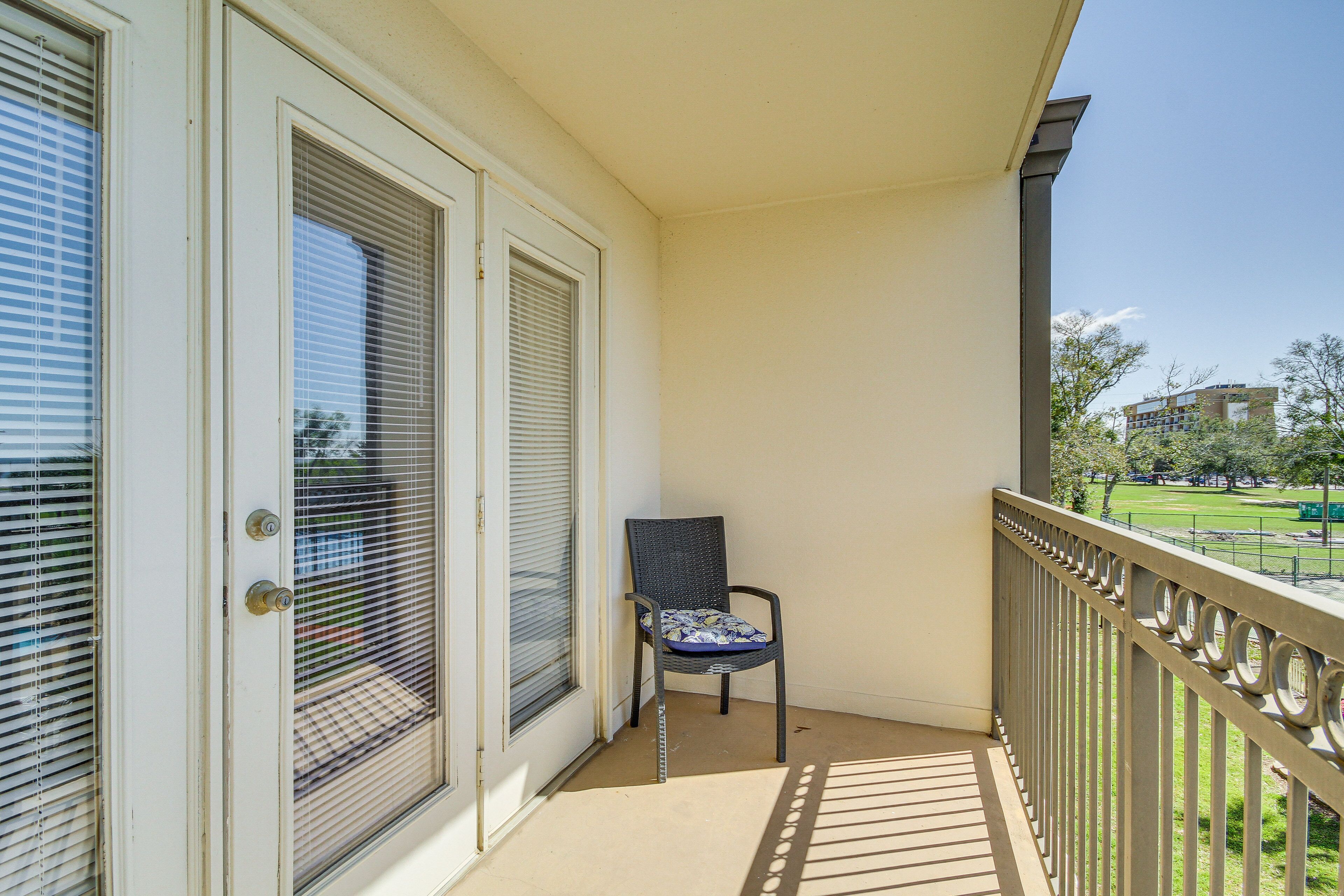 Un balcon avec une chaise, une porte avec une fenêtre, et une vue sur un parc et des immeubles.