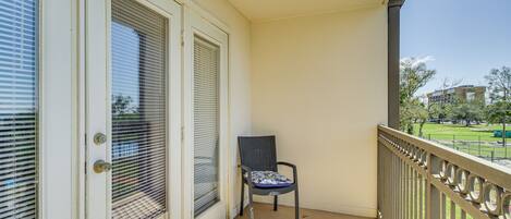 Un balcon avec une chaise, une porte avec une fenêtre, et une vue sur un parc et des immeubles.