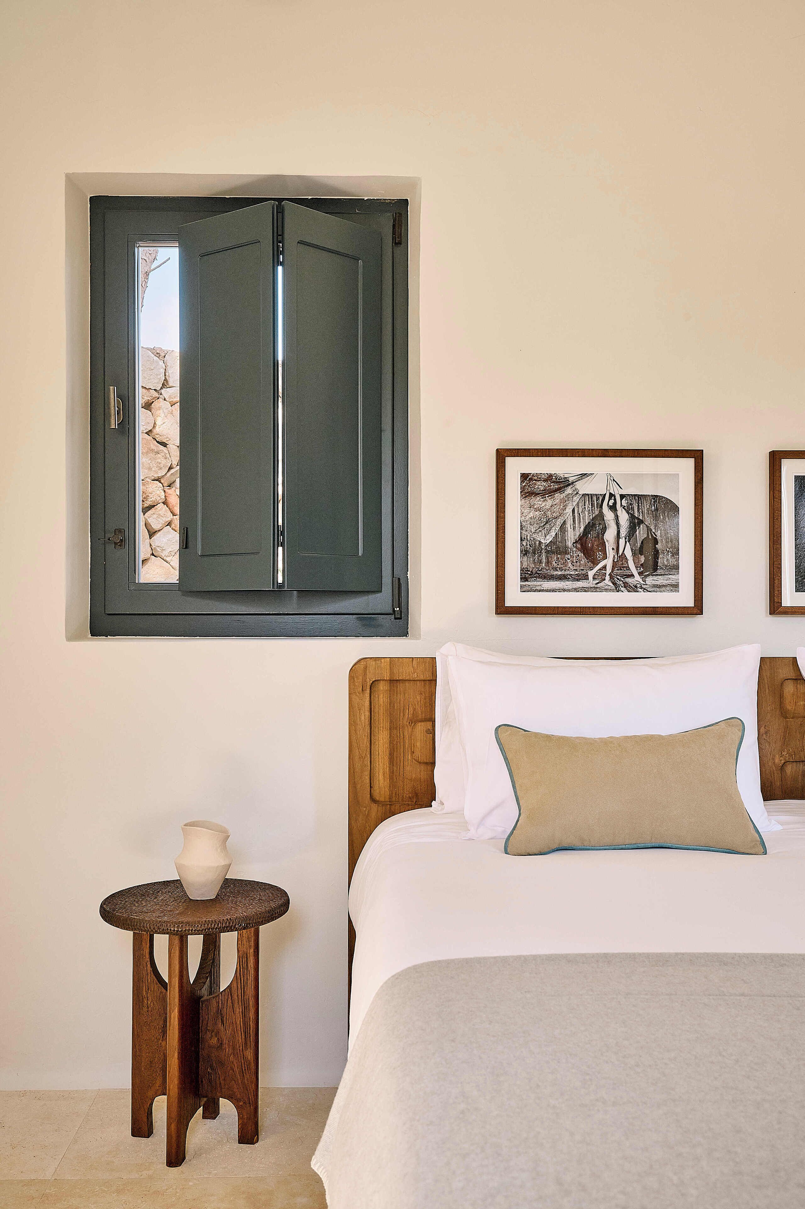 Ein Schlafzimmer mit einem Bett, einem kleinen hölzernen Hocker, einem Fenster mit Blick auf Felsen und einem gerahmten Bild an der Wand.