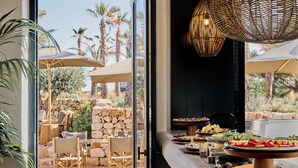 An outdoor dining area with a stone wall, wooden furniture, and a variety of food and drinks on a buffet table. - Hotel Cap Menorca - Relais & Châteaux (Alayor)