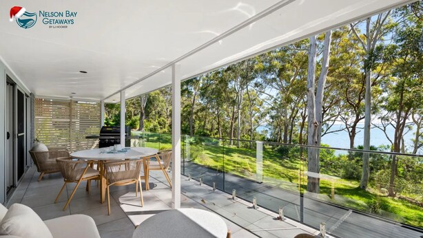 A modern outdoor dining area with a view of the sea and trees. - Tree Tops Water Front Soldiers Point- First Floor (Soldiers Point)