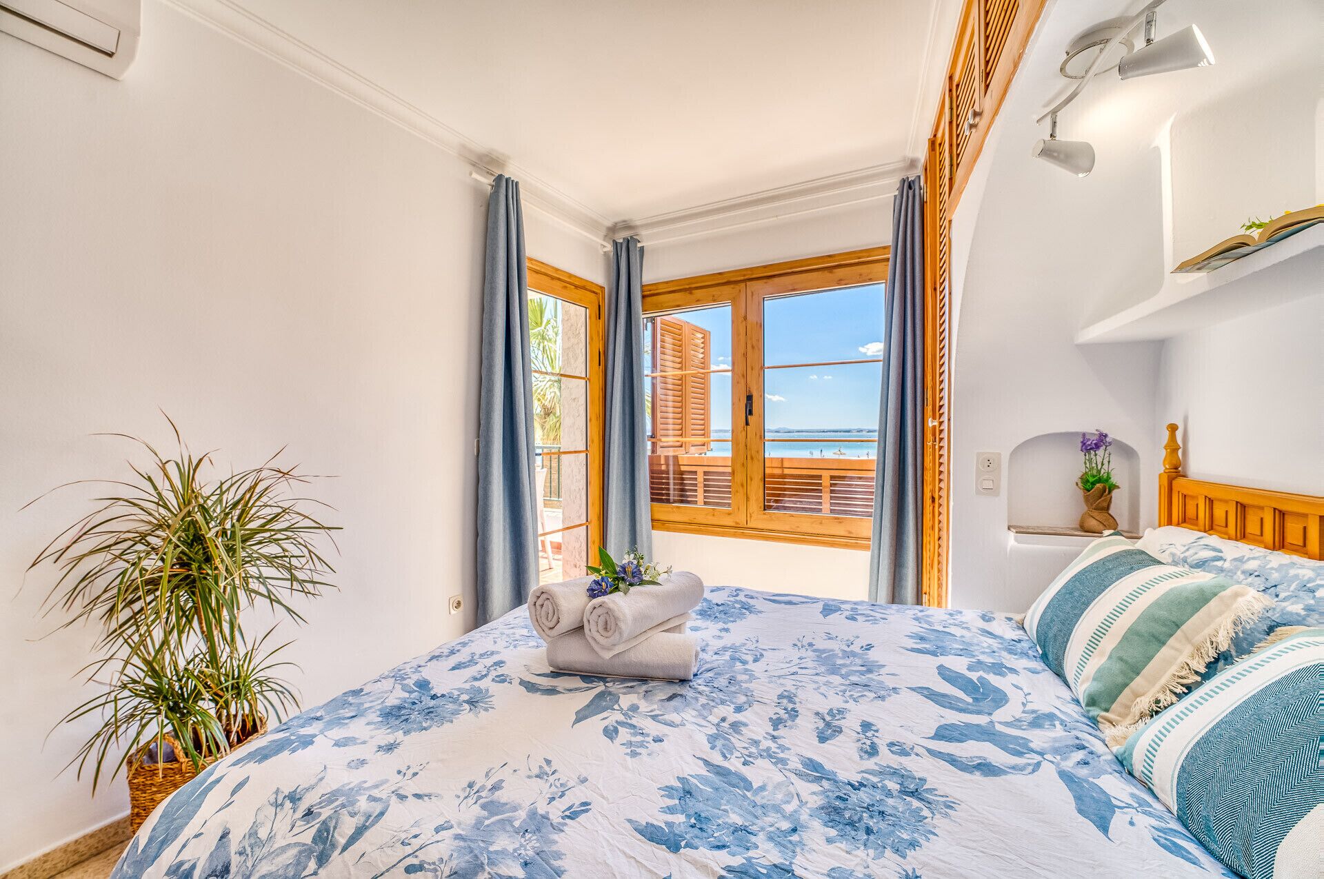 A bedroom with a bed, a potted plant, a window with a view of the sea, and a shelf with a vase.
