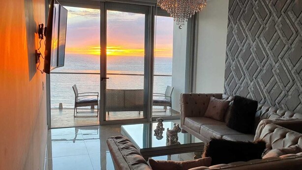 Una sala moderna con una mesa de centro de vidrio, un sofá y un sillón orientados hacia un ventanal con vista al atardecer sobre el mar.