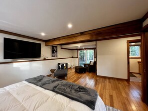 Quarto com uma cama grande, televisão fixada na parede, teto de madeira e porta de correr para uma varanda com vista para árvores.