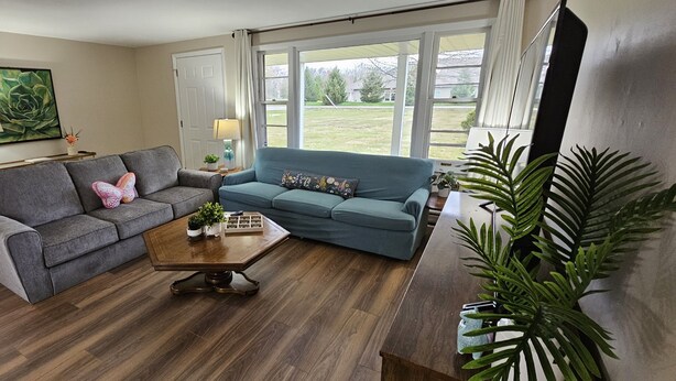 A living room with a grey sofa, a blue sofa, a wooden coffee table, and a large window with a view of a grassy yard. - Escape to a colorful, cozy retreat where families and work crews alike can relax (Fort Wayne)