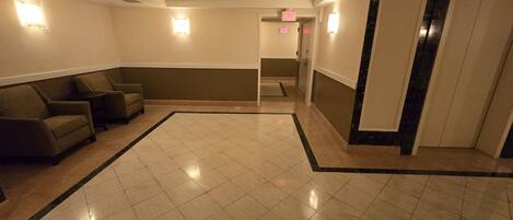 A hotel lobby with a tiled floor, two armchairs, a small table, and a door with an exit sign.
