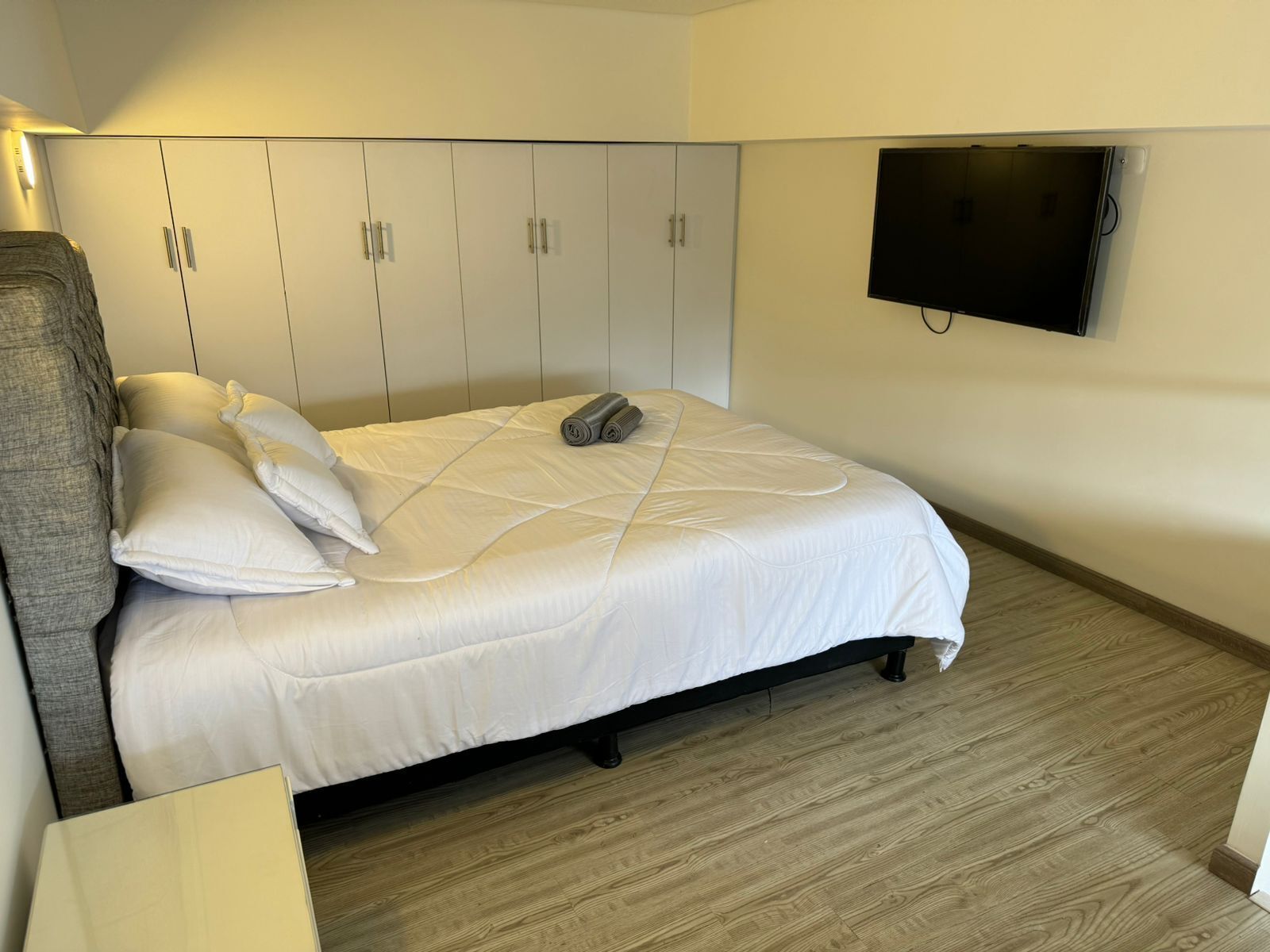 Een keurig opgemaakt bed met witte beddengoed en kussens, een grijs hoofdbord, een flatscreen televisie aan de wand en een houten vloer.