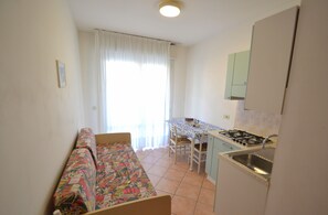 Um quarto pequeno e luminoso com cozinha compacta, uma cama com manta colorida e uma janela com cortinas brancas.