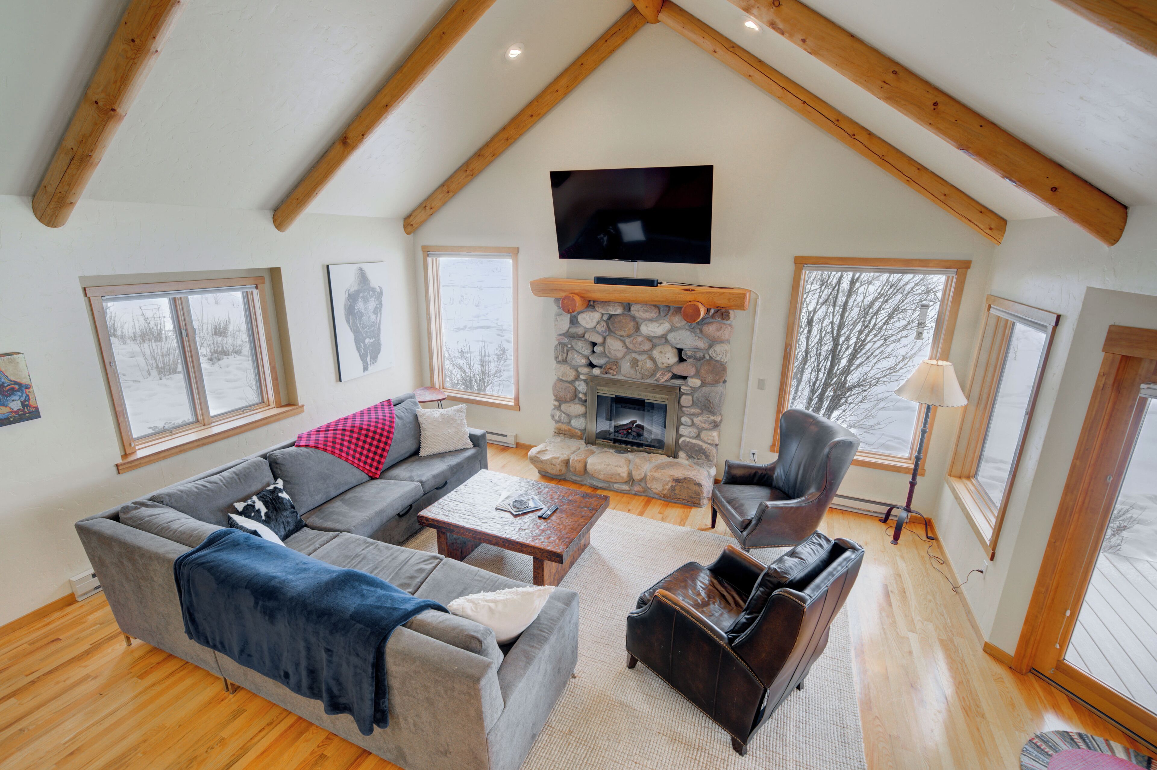 A cozy living room with a stone fireplace, a large TV mounted on the ceiling, and a comfortable seating area with a sofa and armchairs.