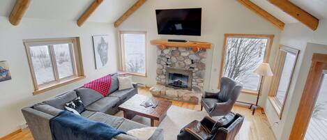 A cozy living room with a stone fireplace, a large TV mounted on the ceiling, and a comfortable seating area with a sofa and armchairs.