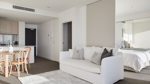 A modern living room with a white sofa, a dining area with wooden chairs, a kitchen in the background, and a large bed with white bedding.