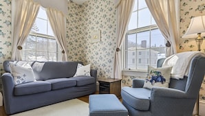 A living room with a grey sofa, a matching armchair, and a small ottoman. There are two windows with curtains, a lamp, and a floral wallpaper. - Peaceful Maine B&B Room  Cozy Stay + Farm-Fresh Breakfast Room 5 (Limerick)