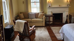 A cozy living room with a beige sofa, a wooden rocking chair, a fireplace, and a large window with curtains.