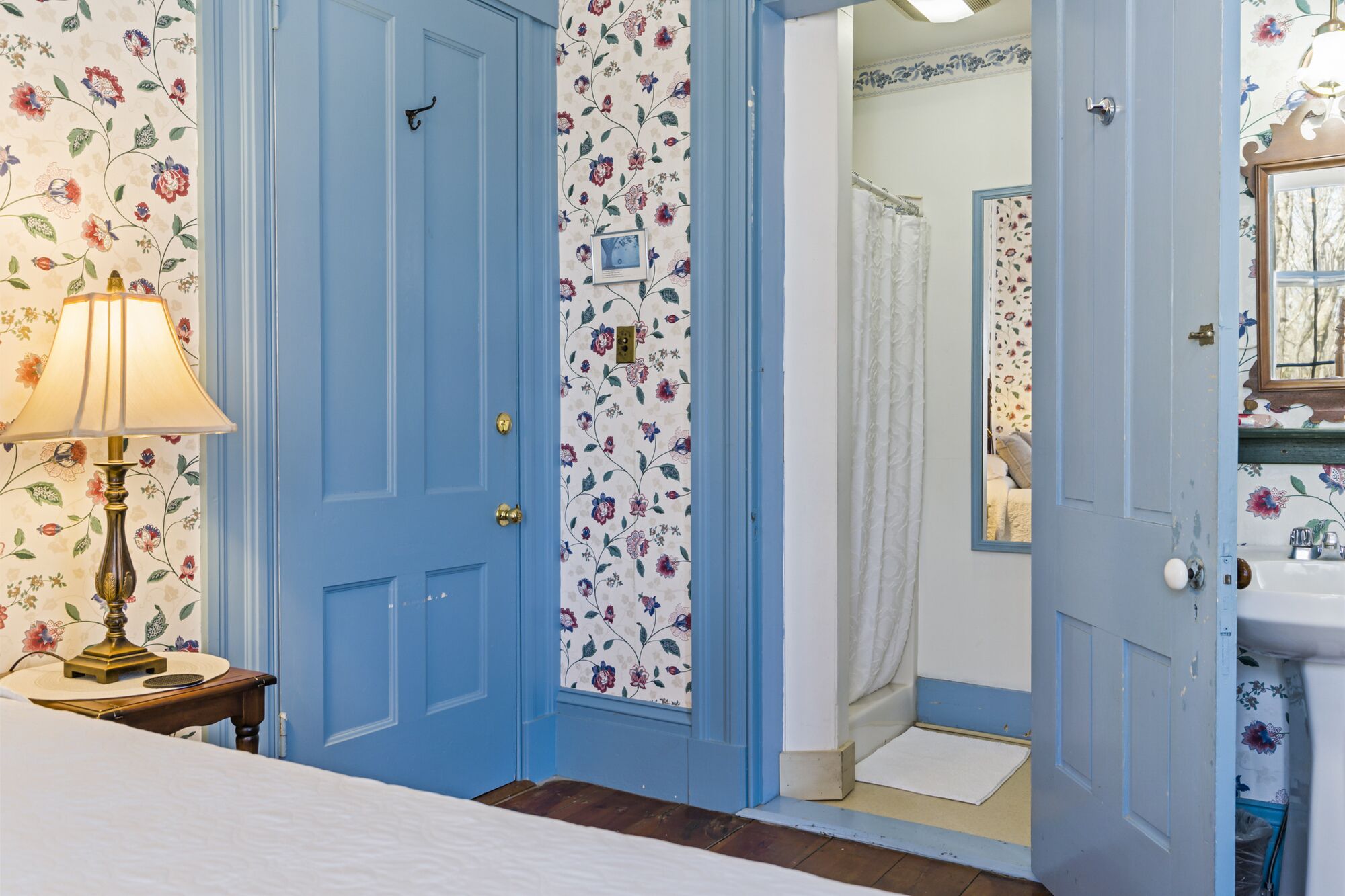 Une chambre à coucher avec un lit, une table de chevet, une lampe, une porte recouverte d’un papier peint à motifs floraux, et une salle de bain visible à travers une porte ouverte.