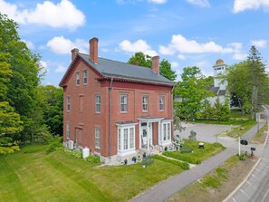 A red brick building with a white porch, surrounded by greenery and a clear sky.