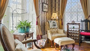 Une pièce avec un papier peint à motifs, une chaise, une table avec une lampe, une plante en pot et une fenêtre avec des rideaux.