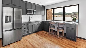 A modern kitchen with grey cabinets, stainless steel appliances, and a large window offering a view of a brick building and hills. - Luxury King Studio Downtown Main Street (Hill City)