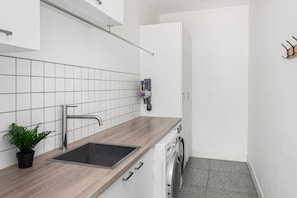 A modern kitchen with a sink, a plant, a washing machine, and a door leading to another room.
