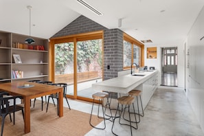 A modern kitchen with a large island, a dining area with a wooden table and chairs, and a bookshelf with books. - The Atrium Abbotsford House BBQ and Parking (Abbotsford)
