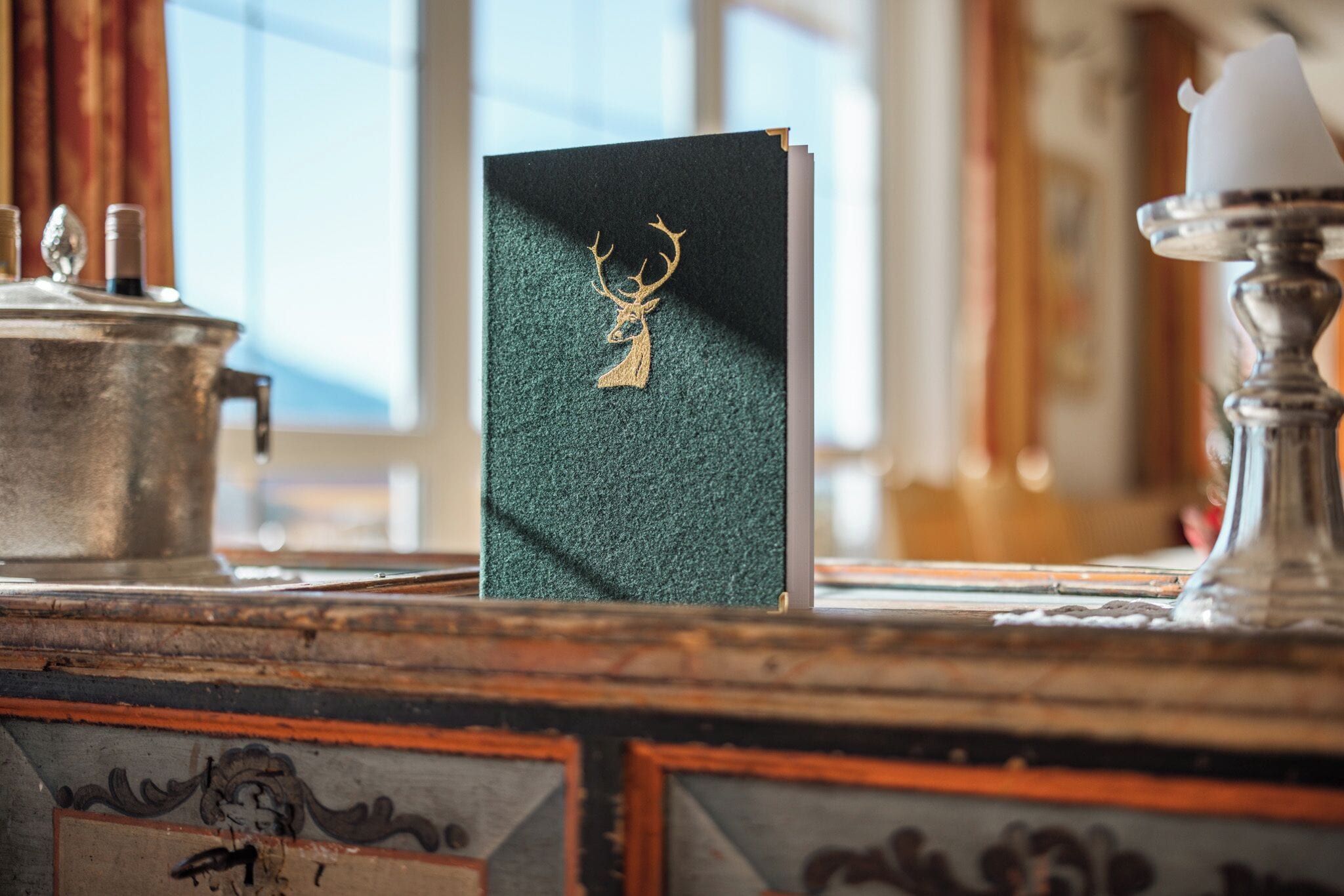 Un libro con un emblema de ciervo sobre una superficie de madera, posiblemente un escritorio, y un candelabro cerca.