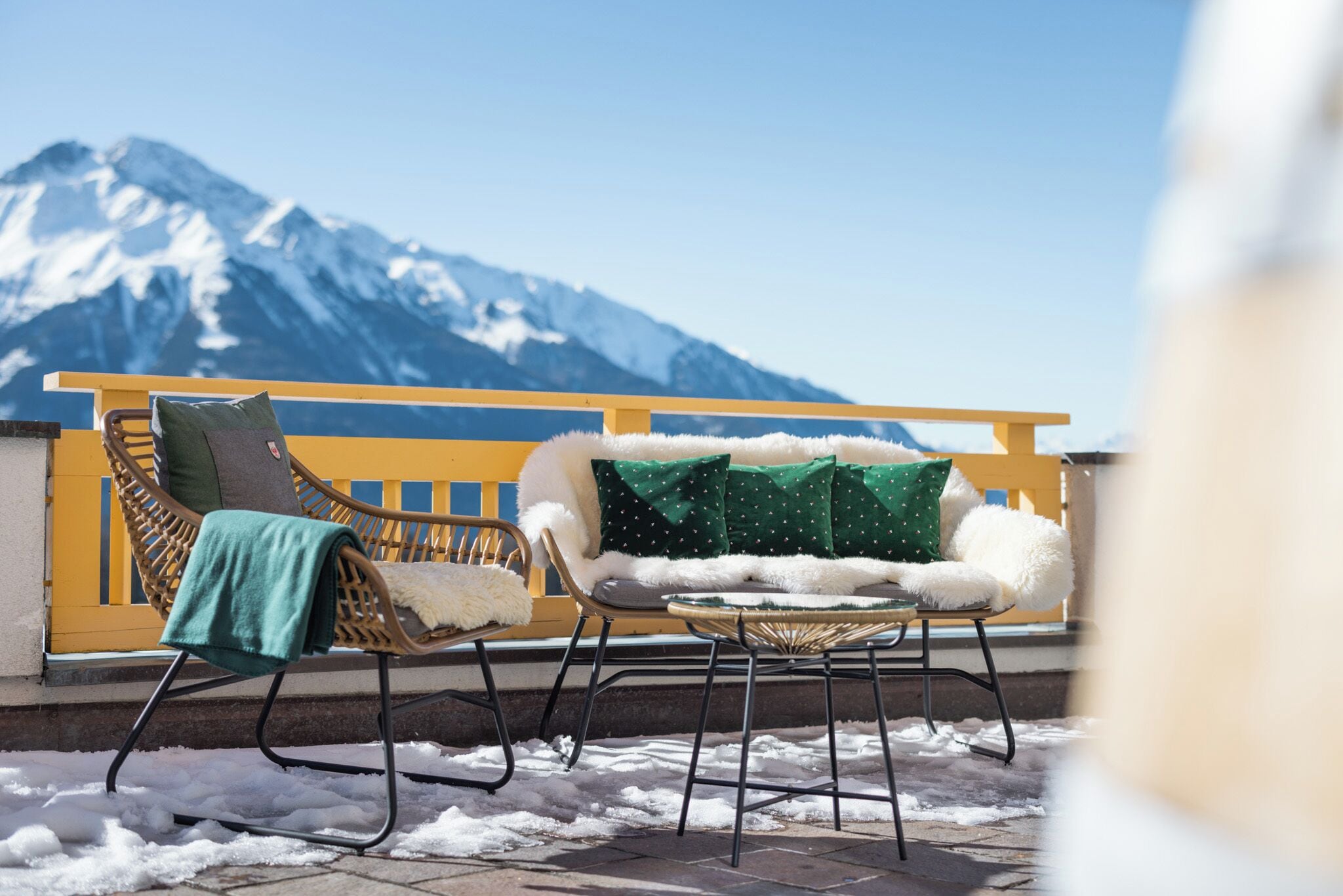 Ein Balkon mit gelber Brüstung, zwei Korbstühlen und einem weißen, mit Fell bezogenen Sofa.